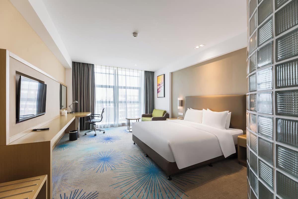 A hotel room with a large bed, a desk with a computer, a TV, and a window with curtains.