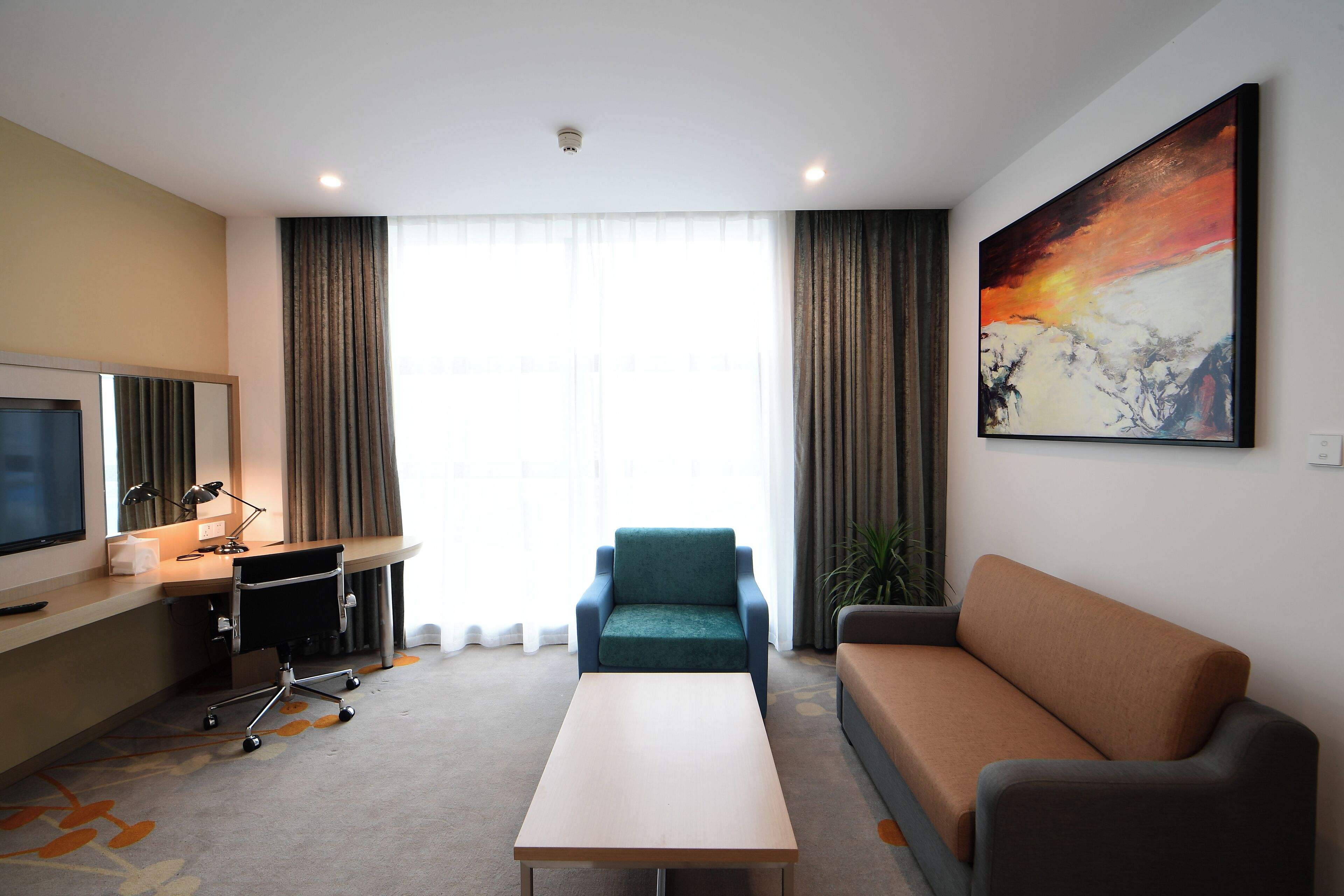 A hotel room with a flat-screen TV, a desk with a lamp, a green armchair, a brown sofa, and a large abstract painting on the wall.