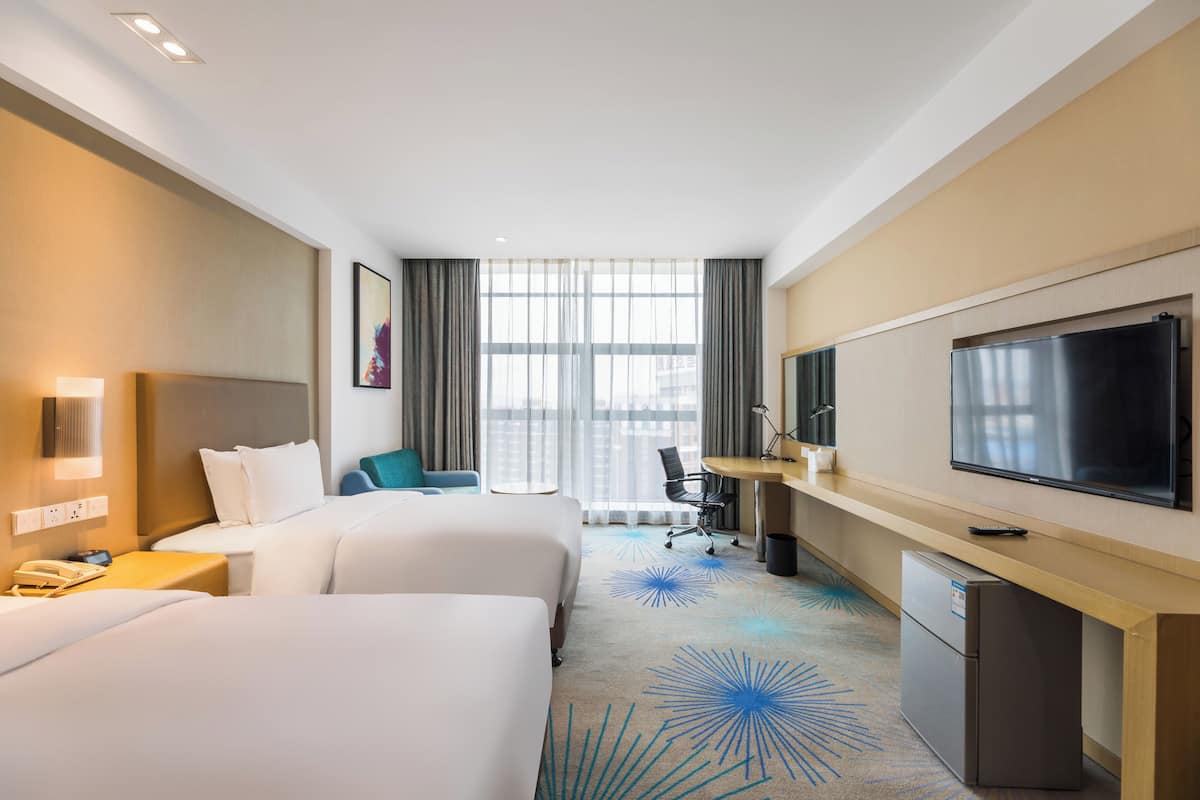 A hotel room with two beds, a desk, a television, and a window with curtains.