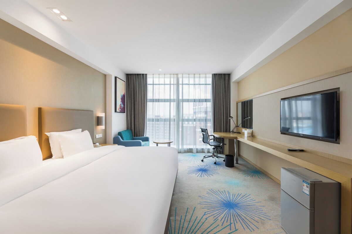 A hotel room with a large bed, a desk with a chair, a television, and a window with curtains.
