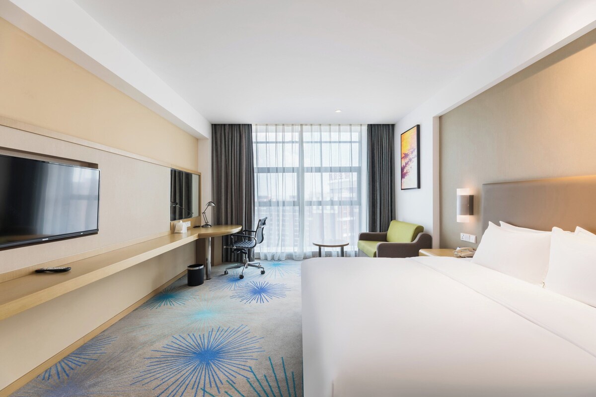 A hotel room with a large bed, a desk with a chair, a television, and a window with curtains.