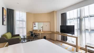Una habitación de hotel moderna con una cama grande, un escritorio con silla, televisión y ventanas amplias con cortinas.