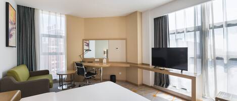 A hotel room with a large bed, a desk, a chair, a television, and large windows with curtains.