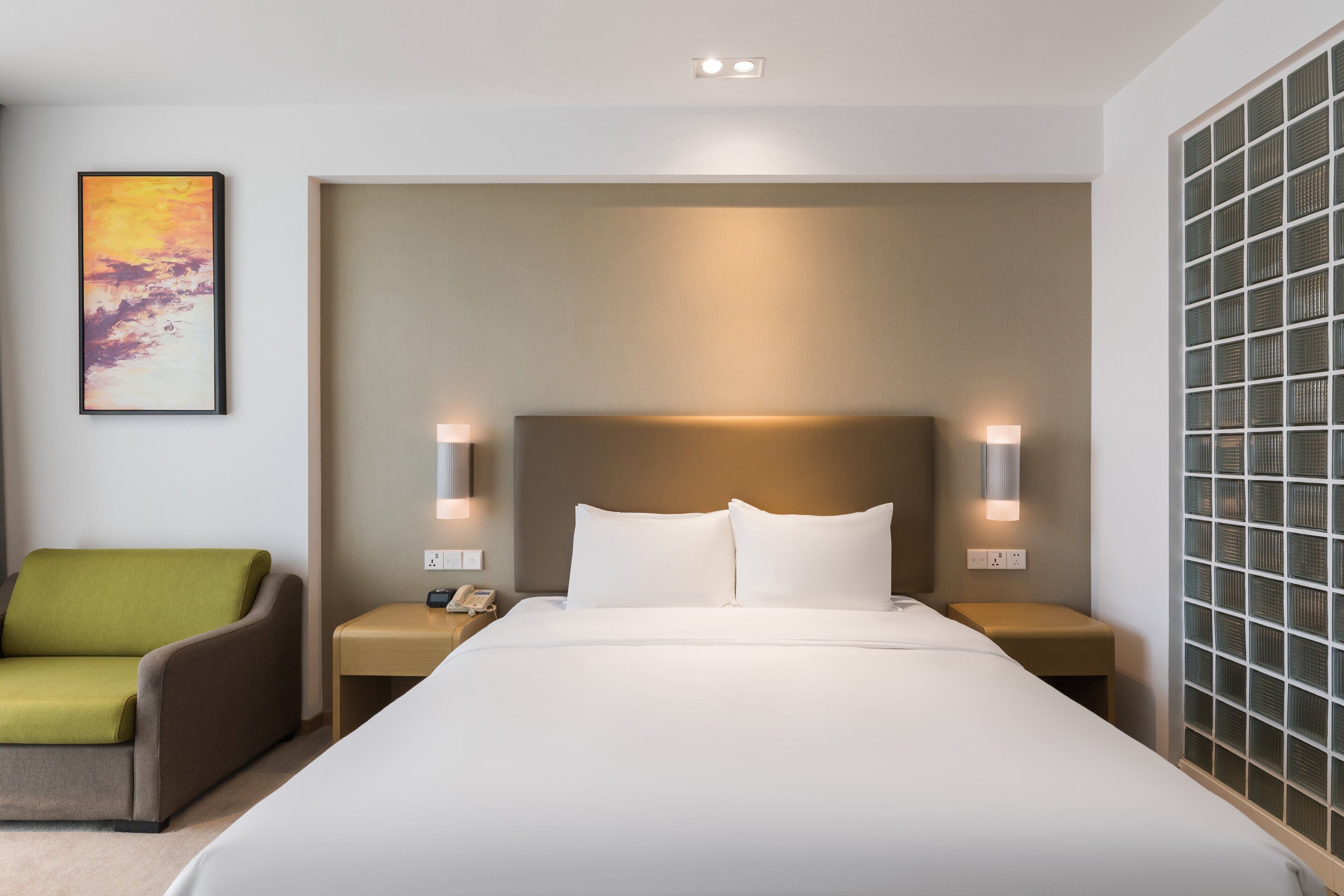 A modern hotel room with a large bed, a green armchair, bedside lamps, and a wall-mounted artwork.