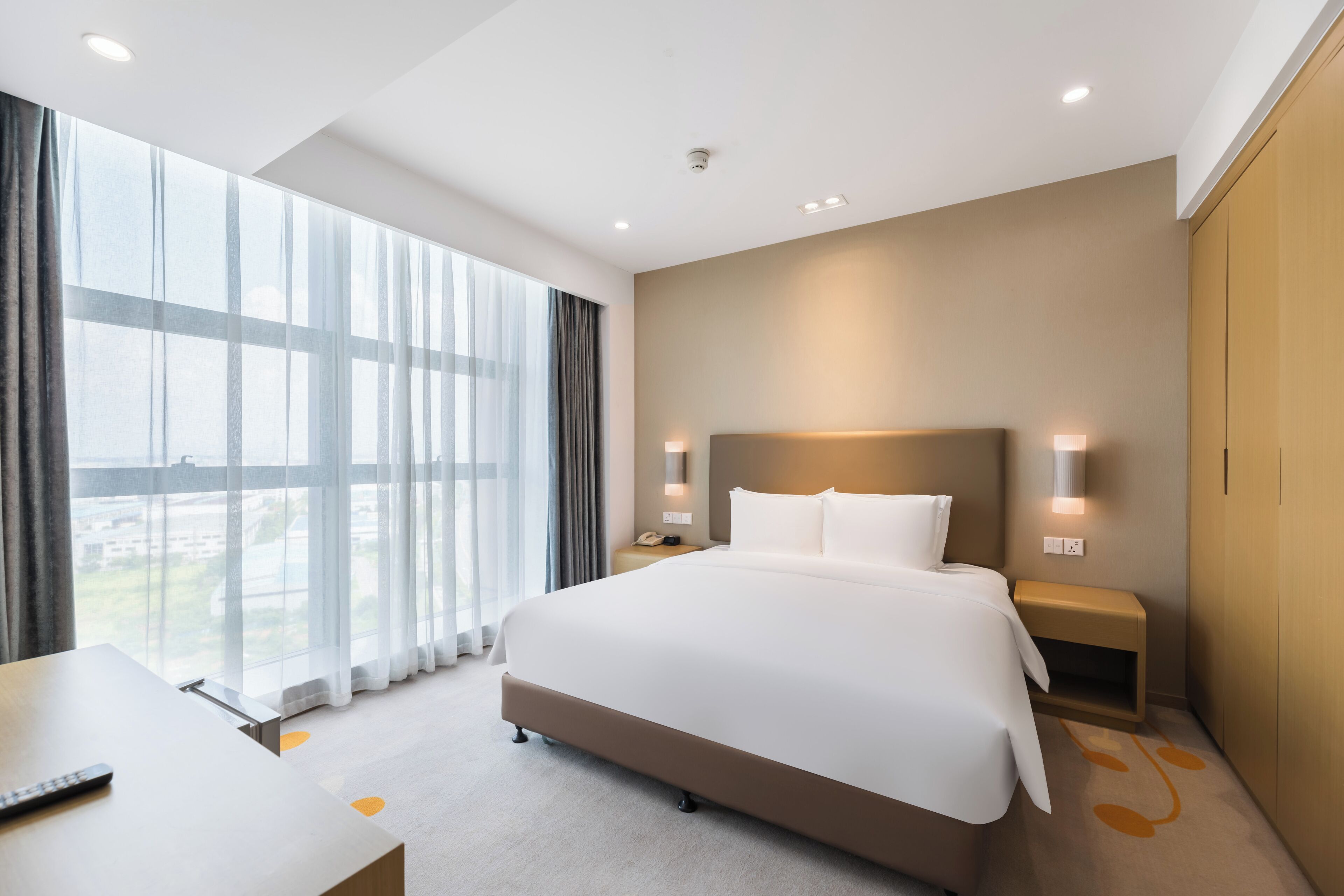 A modern hotel room with a large bed, bedside tables, a desk, and a view of the city.