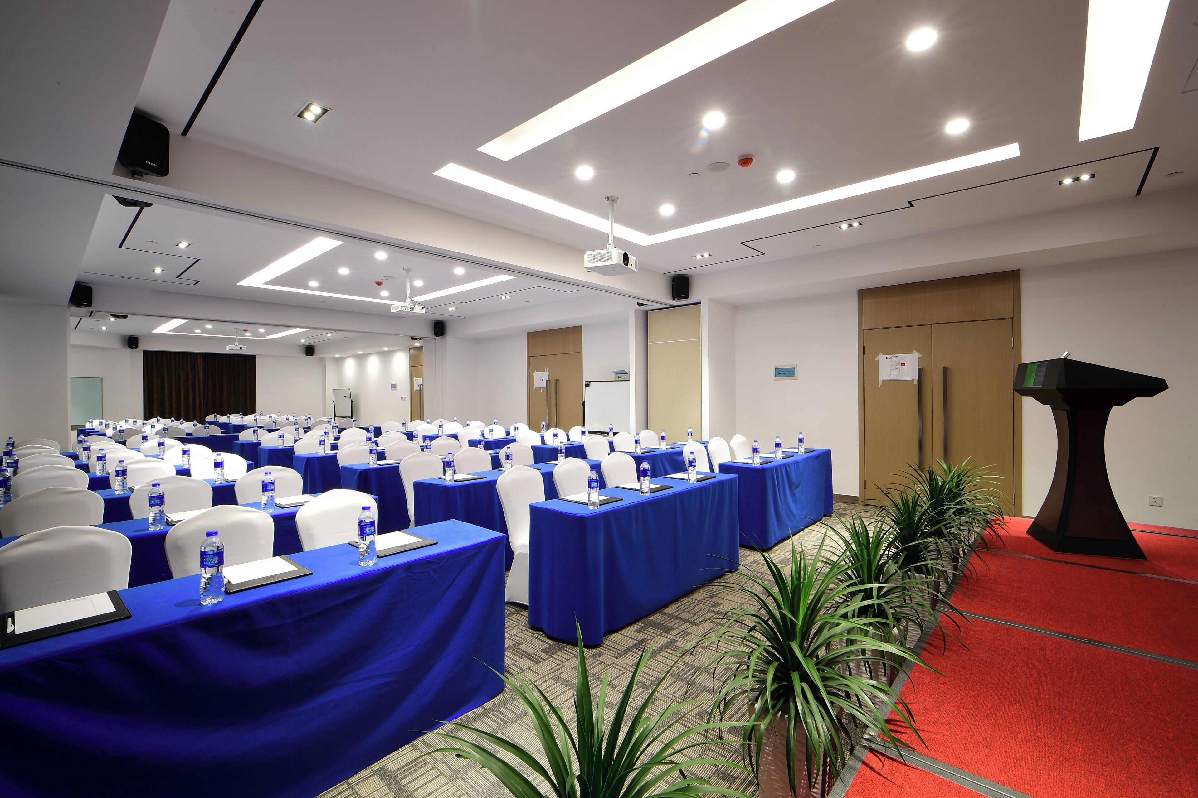 A conference room with rows of tables, chairs, and water bottles, equipped with a projector and a podium.