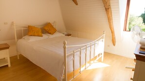 Un dormitorio con estructura de cama metálica blanca, dos almohadas amarillas, suelo de madera y ventana con vistas a la vegetación.