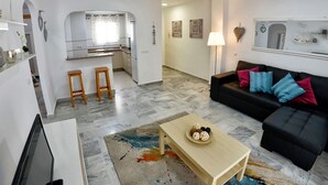 Een moderne woonkamer met een zwarte bank, een houten salontafel en een televisie. Er is een keukenhoek met een spoelbak en barkrukken.