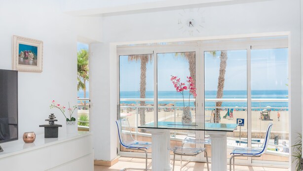 A bright room with a glass table and chairs, a TV, and a view of the beach.