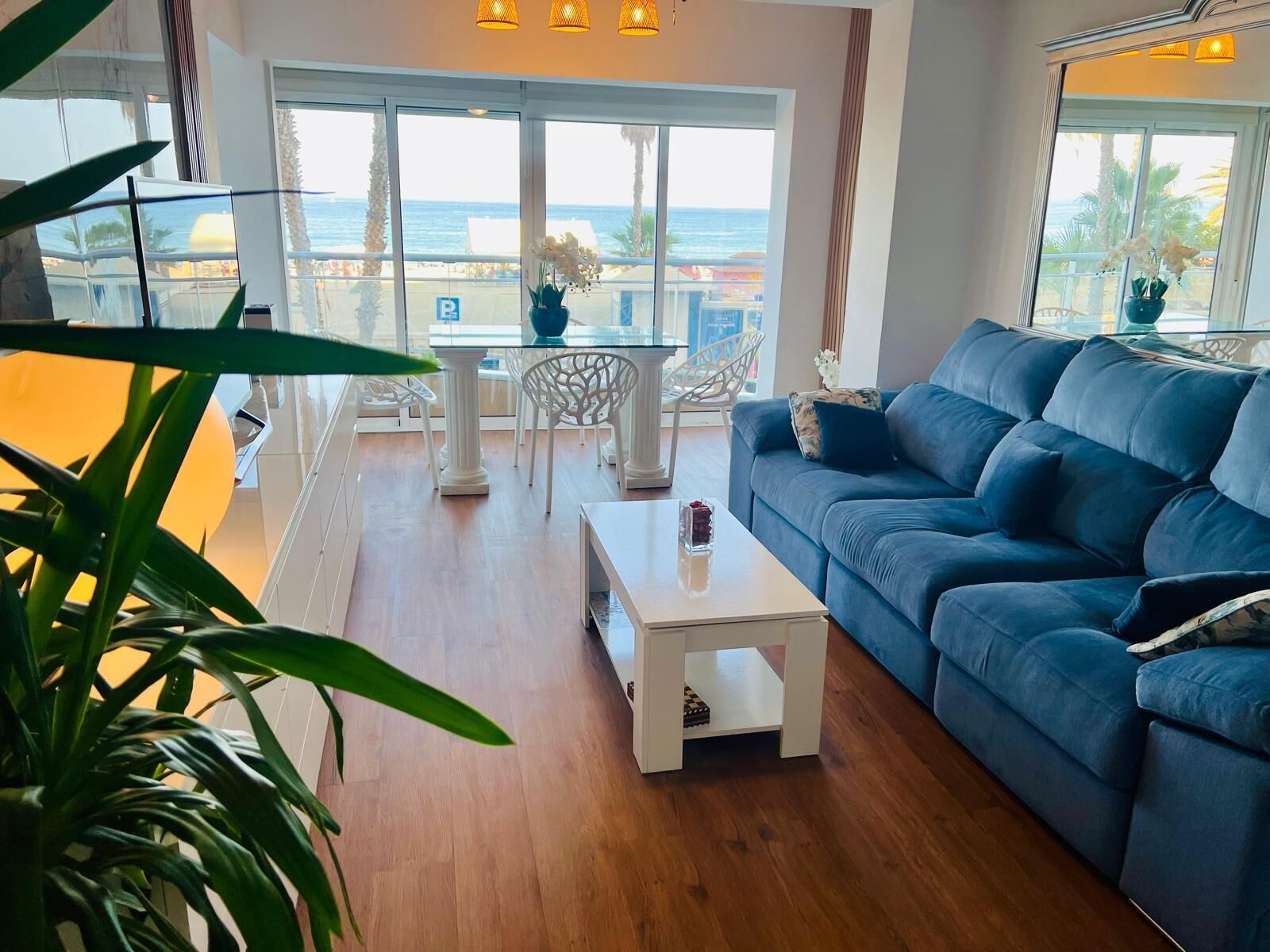 Una sala de estar moderna con un sofá azul, una mesa de centro blanca y una mesa de vidrio con sillas. Ventanas amplias ofrecen vista a la playa y palmeras.