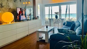 A modern living room with a television, white cabinets, a glass coffee table, a blue sofa, and a balcony with a view of the sea.