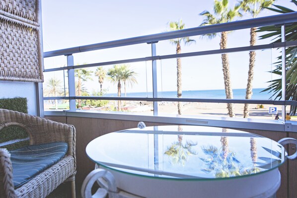 A balcony with wicker furniture, a glass-top table, and a view of the beach and palm trees.