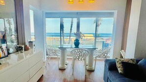 A modern living room with a glass table and chairs, a sofa, and a view of the beach.