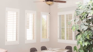 Comedor con una mesa redonda, seis sillas, ventilador de techo y un ventanal con contraventanas.