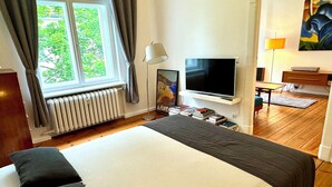 Ett sovrum med en stor säng, en tv, ett element, ett fönster med gardiner och en tavla på väggen.