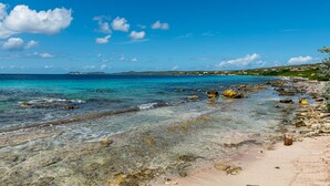 A beach with clear water, rocks, and a distant view of houses. - Tropical oceanfront villa, full AC, private pool, great diving and snorkeling (Kralendijk)