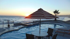 Ein Poolbereich mit Tisch und Stühlen, einem gestreiften Sonnenschirm und Blick auf den Ozean bei Sonnenuntergang.