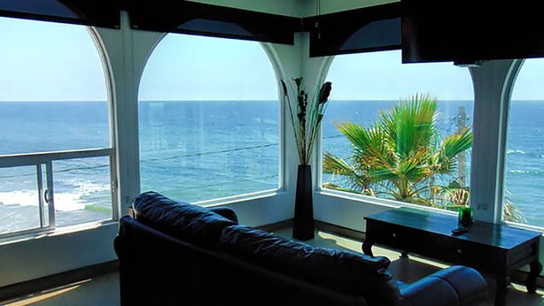 Ein Zimmer mit großem Fenster mit Blick auf den Ozean, einem schwarzen Sofa, einem kleinen Tisch und einer Pflanzvase.