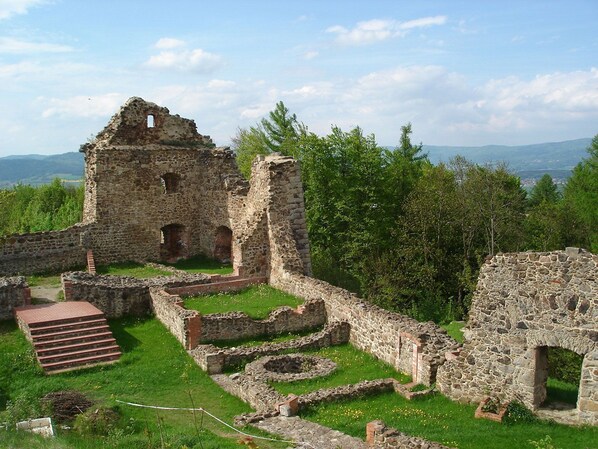 Restes d'una construcció de pedra amb una escala i murs circumdants, en una zona verda amb arbres i muntanyes al capdamunt.