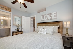 Une chambre avec un grand lit, une tête de lit en bois, un panneau mural sur lequel est écrit le mot « AMOUR », un ventilateur de plafond et une table de chevet avec une lampe.