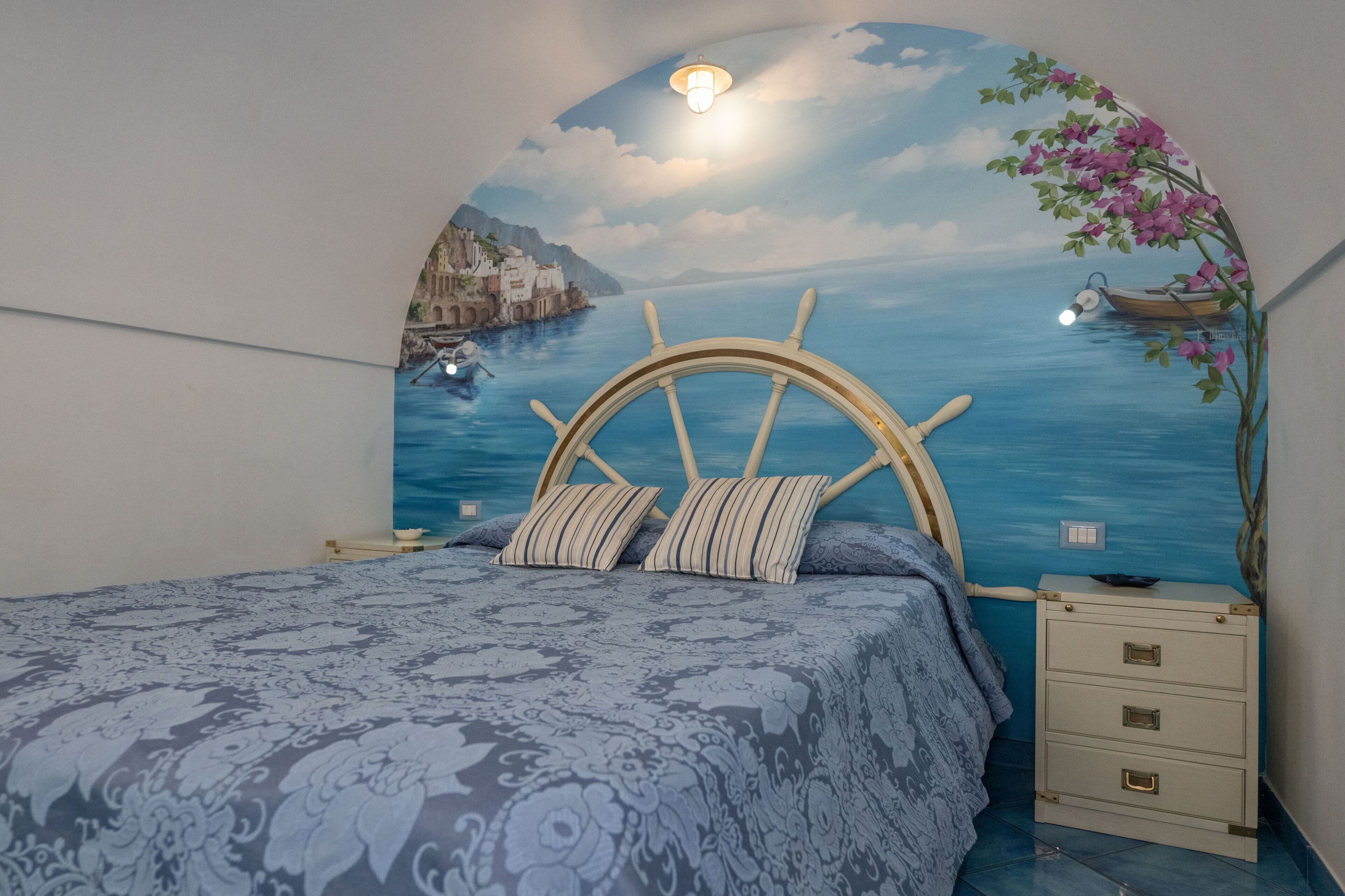 A bedroom with a bed, a nightstand, a dresser, and a mural of a coastal scene.