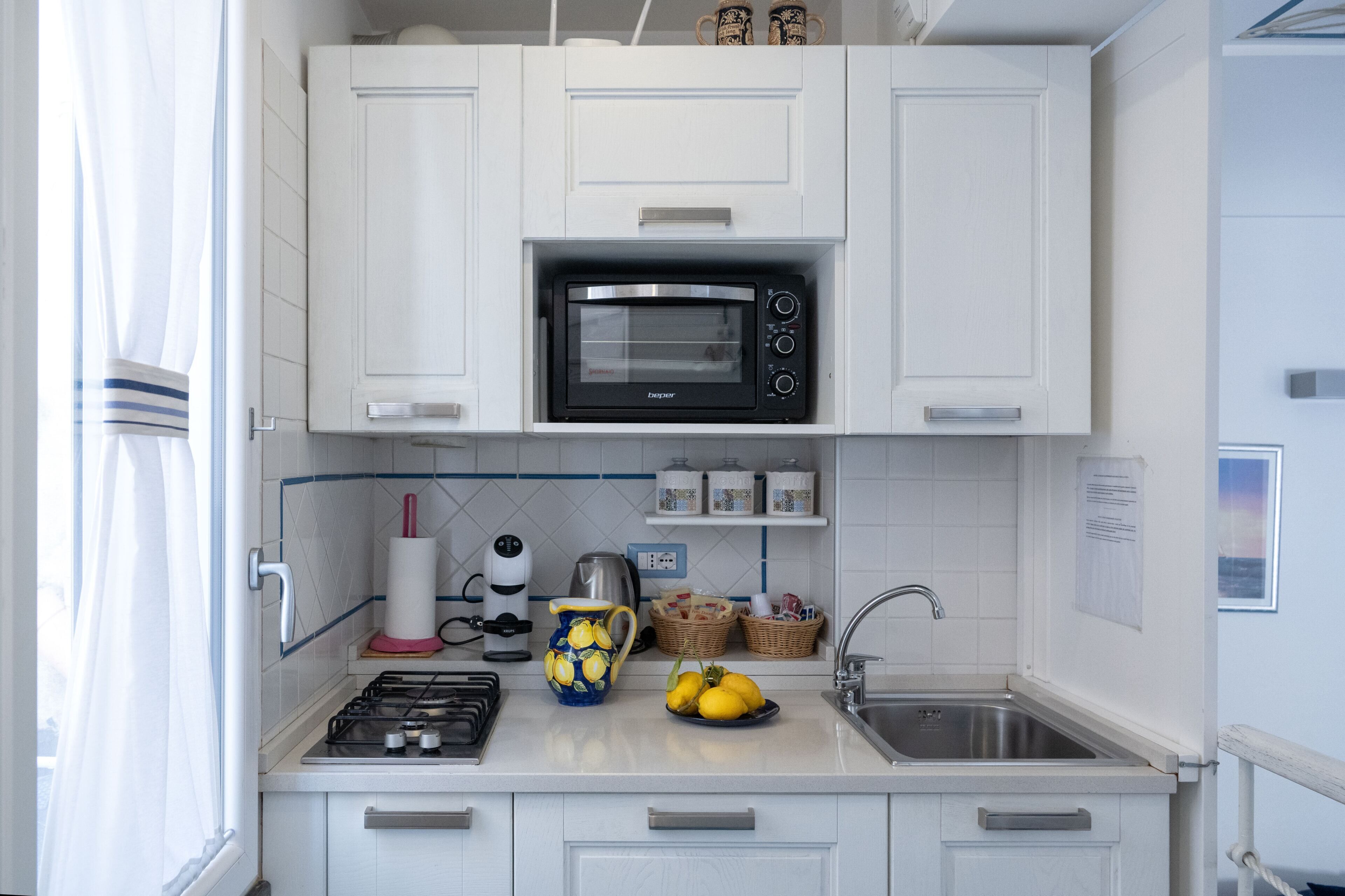 Una cocina compacta con gabinetes blancos, microondas integrado, estufa a gas, un lavabo y una ventana con cortinas.
