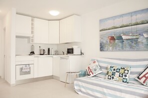 A compact room with a kitchenette, a bed with striped bedding, and a painting of boats on the wall.