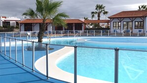 Une piscine au design incurvé, entourée d’une rambarde métallique. On aperçoit des palmiers et un bâtiment avec un toit recouvert de tuiles rouges à l’arrière-plan.