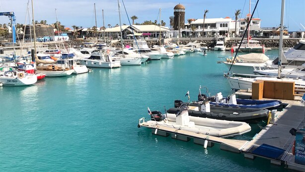 Un port de plaisance où sont amarrés différents types de bateaux, une tour à eau et un bâtiment sur lequel on peut lire l’inscription « Chocolaterie ».