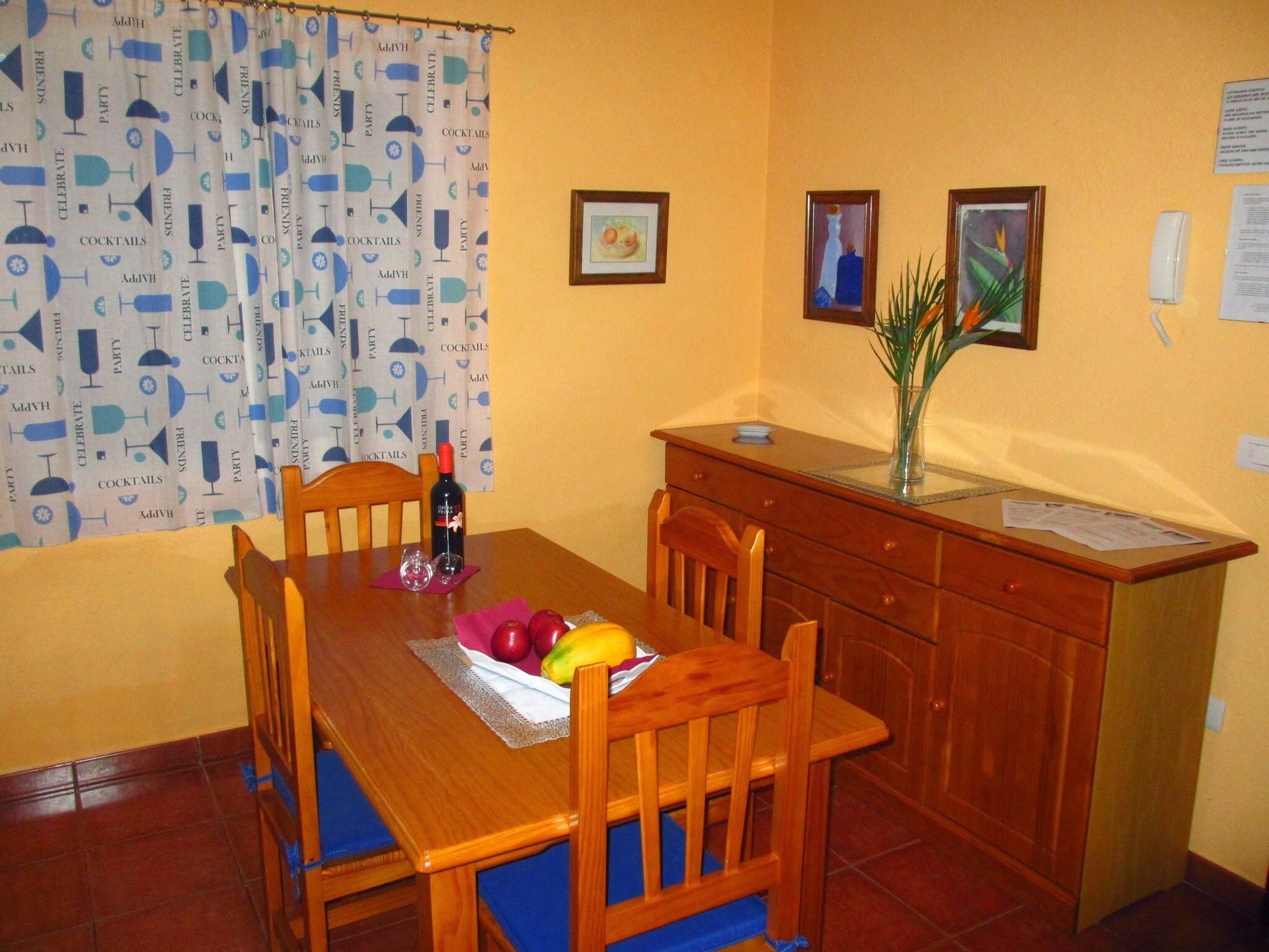 A dining area with a wooden table, chairs, a bottle of wine, and a fruit bowl.