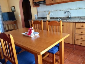 A dining area with a wooden table and chairs, a kitchen with a sink and cabinets, and a tiled backsplash.