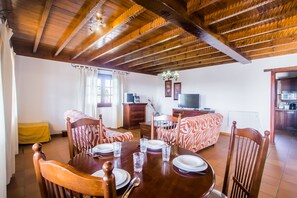 A dining area with a wooden table set for six, surrounded by patterned chairs. A television is mounted on the wall, and a kitchen area is visible in the background.