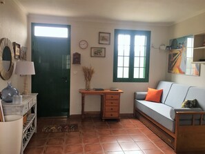 A room with a green door, a desk with a lamp, a sofa with an orange cushion, and a window with green shutters.