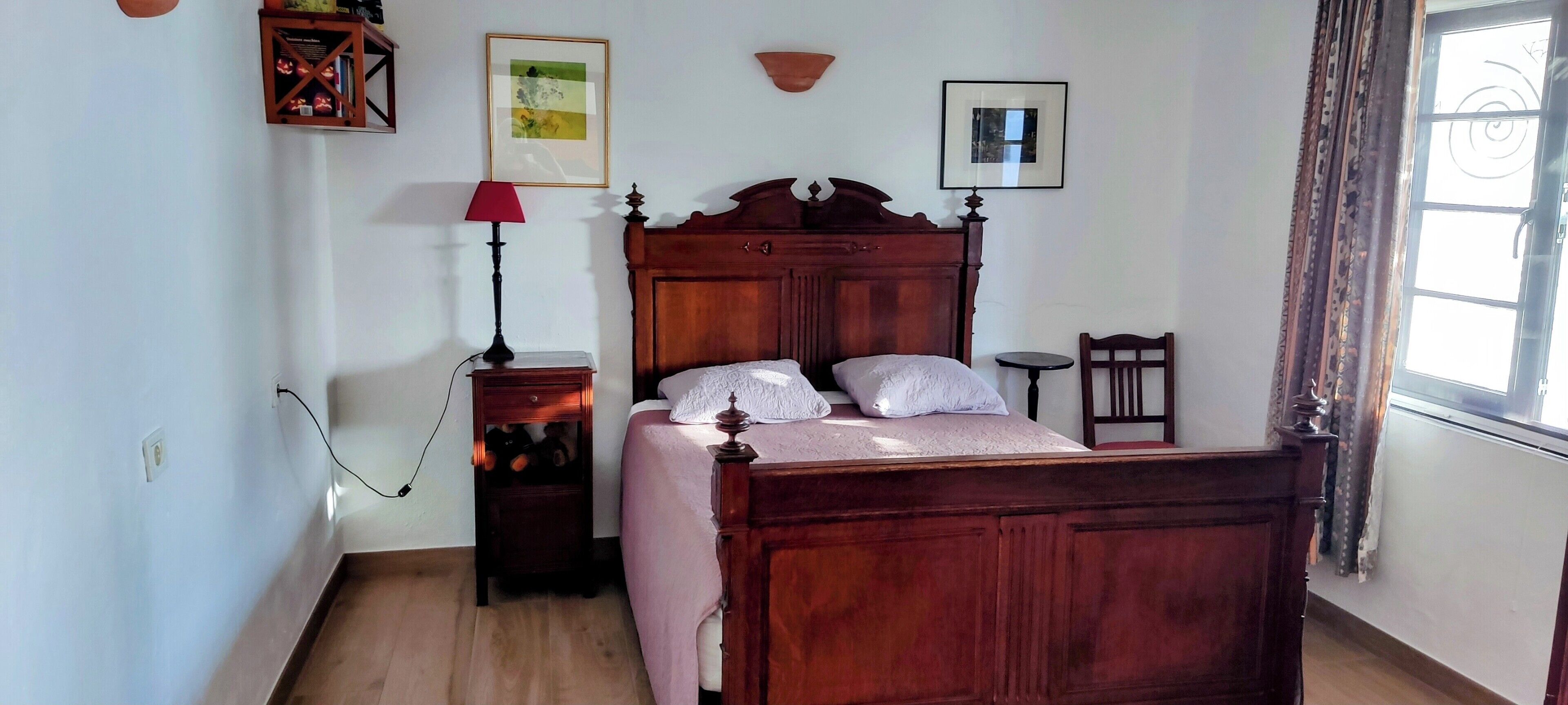 A bedroom with a wooden bed, a bedside table, a lamp, a small table, and a window with curtains.