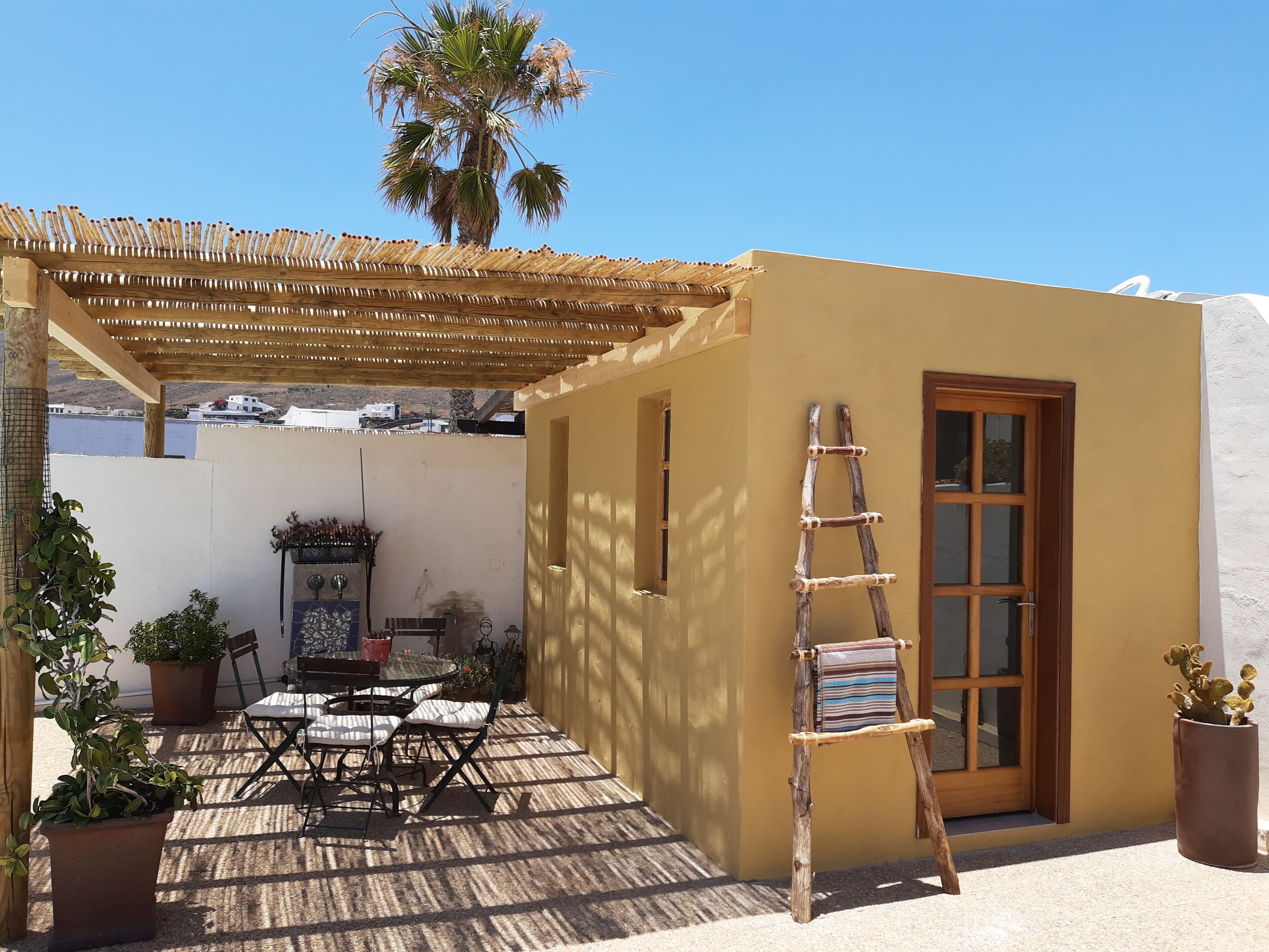 A patio with a wooden pergola, a table with chairs, potted plants, and a ladder leaning against a wall.