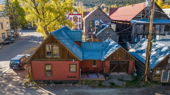 A rustic cabin with a red exterior and a blue roof, surrounded by other wooden buildings and a church in the background.