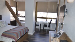 A bedroom with a bed, a desk with a laptop, and a window with a view of the outside.