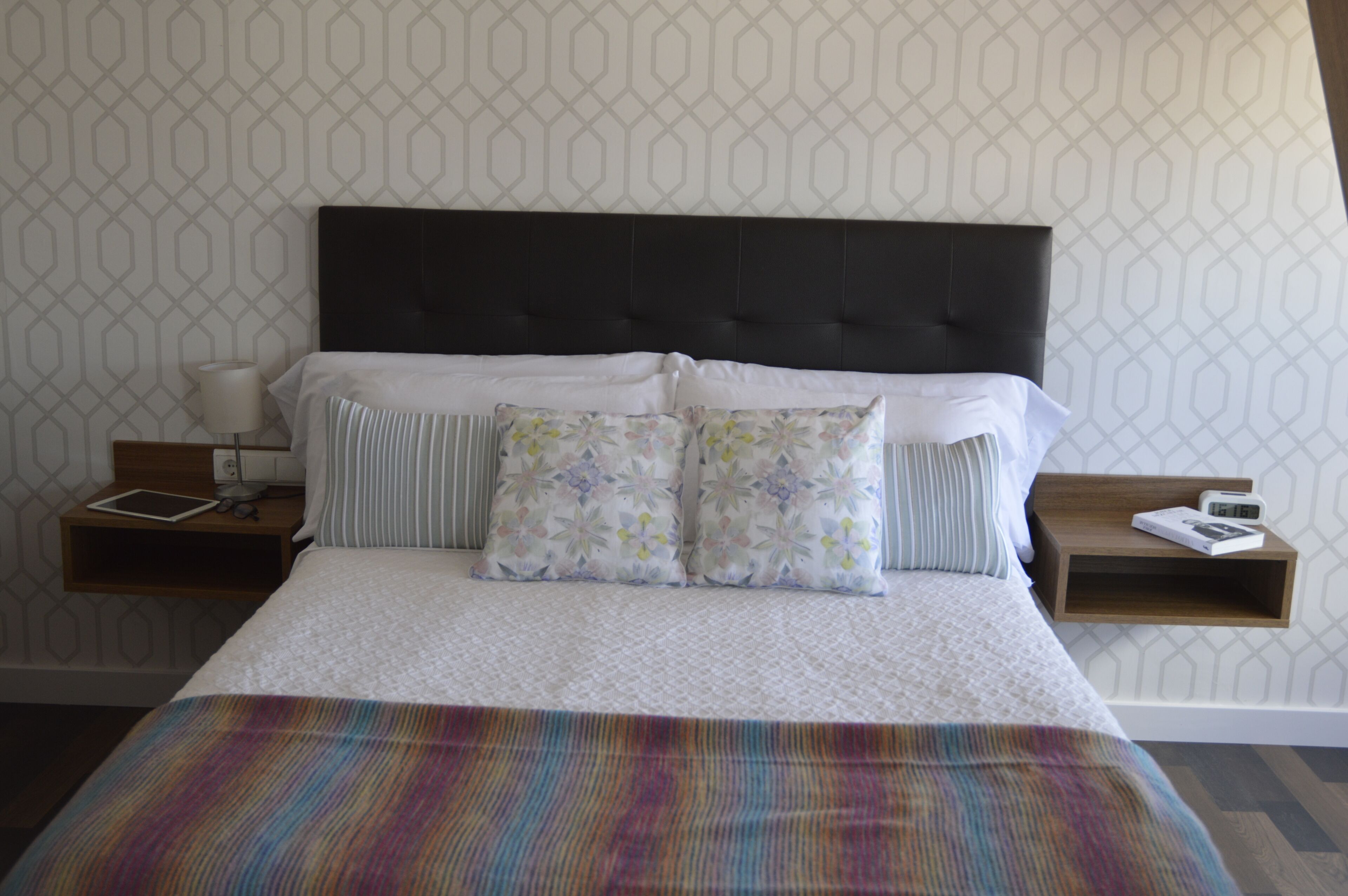 Un lit bien fait, agrémenté d’un coussin à rayures et de deux coussins à motifs floraux, une table de chevet avec une lampe et un livre, et un mur orné d’un motif géométrique.