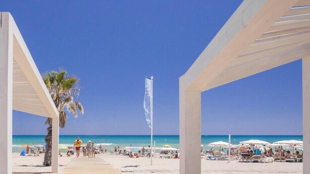 A beach scene with a wooden walkway leading to the shore, palm trees, and beach umbrellas.