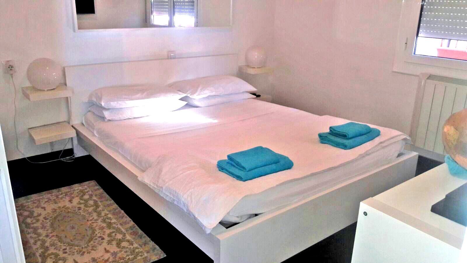 A white bed with two blue towels, a bedside table, a nightstand, a wall-mounted light, and a window with blinds.