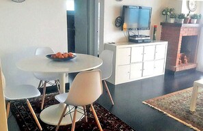 Un coin repas avec une table ronde, des chaises, un saladier de fruits, un téléviseur posé sur un meuble et une cheminée.