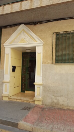 Ein Gebäudeeingang mit gelber Tür, einer kleinen Veranda mit weißem Geländer und einem Fenster mit grünen Fensterläden.