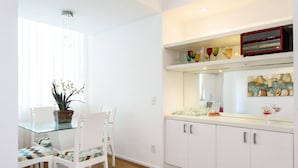 A dining area with a glass table, white chairs, a potted plant, and a kitchen area with white cabinets and a countertop. - All in Rio Conforto e Silencio (Rio de Janeiro)