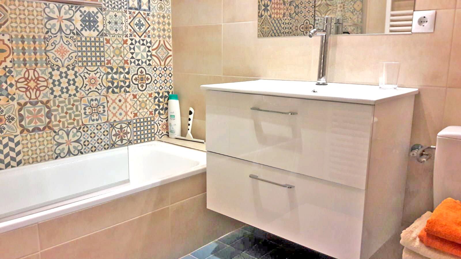 A bathroom with a bathtub, a white vanity with drawers, a mirror, and patterned tiles on the wall.