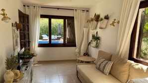 A cozy living room with a sofa, a small round table, and a bookshelf. There is a window with curtains, a guitar, and a plant hanging on the wall. - Villa on the beach in Pals-Begur with barbecue and private pool (Pals)