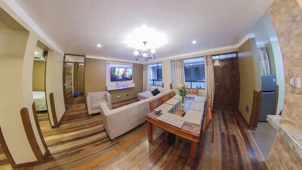 Een ruime woonkamer met een houten vloer, een bank, een eettafel en een aan de muur bevestigde televisie.