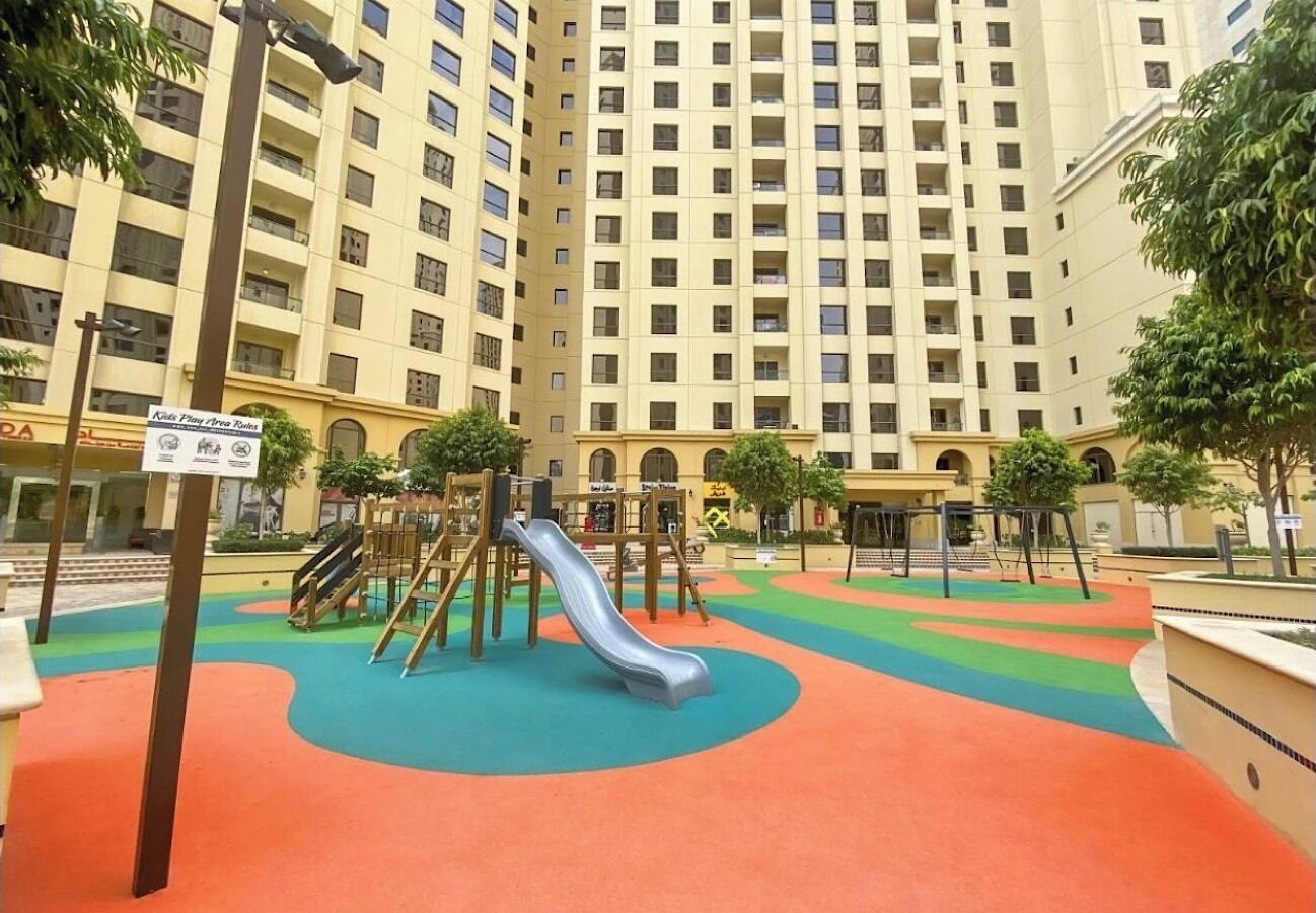 Un terrain de jeux équipé d’un toboggan et de structures d’escalade, entouré d’un immeuble à plusieurs étages doté de balcons.
