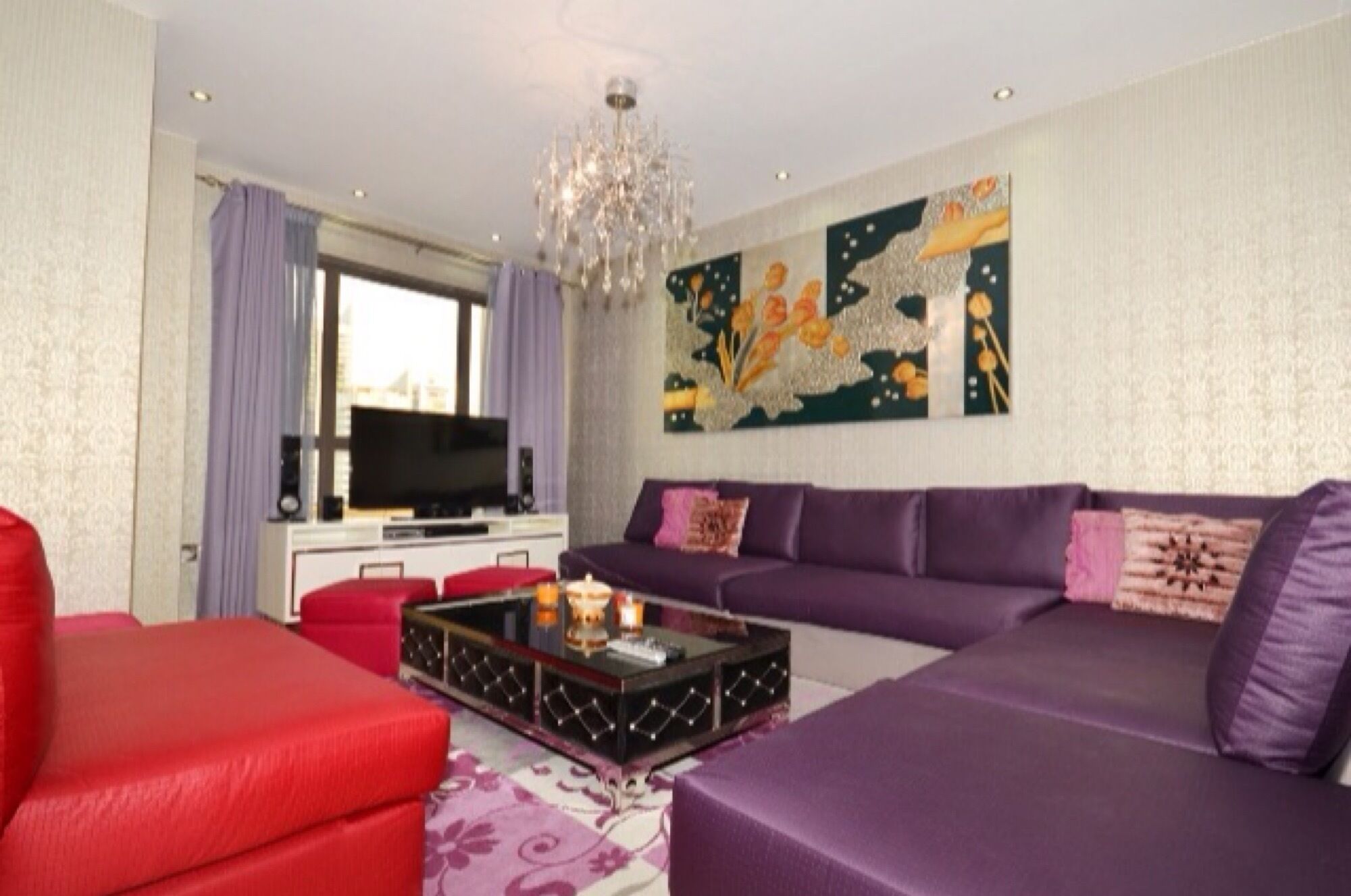 Una sala de estar moderna con un sofá grande, un sillón rojo y una mesa de centro. Hay un televisor sobre un mueble bajo, una araña y una ventana con cortinas.