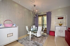 A modern living room with a glass dining table, white chairs, a chandelier, and a view of buildings through large windows.