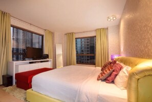 A hotel room with a large bed, a TV on a white cabinet, and a view of tall buildings through the windows.
